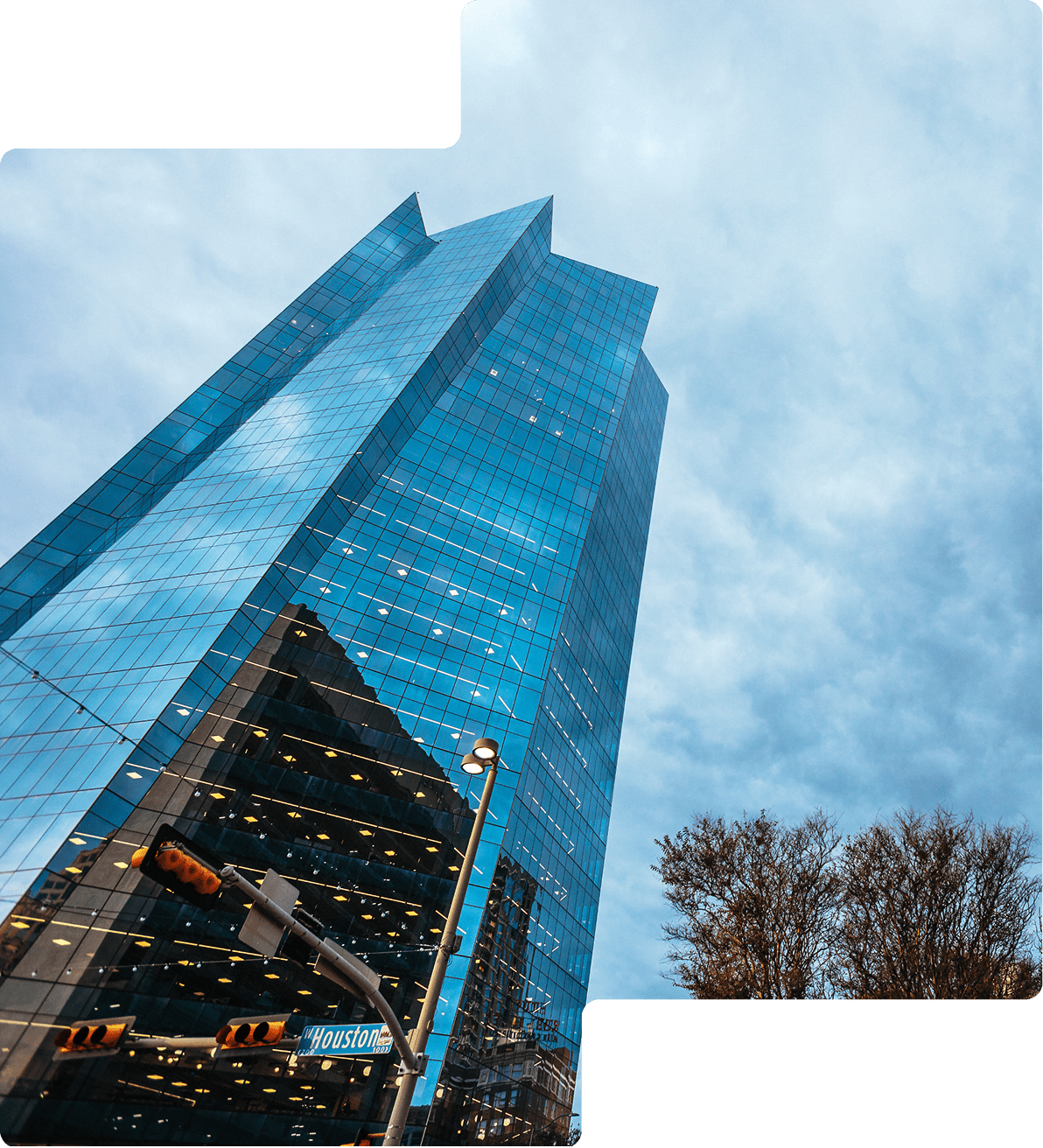 Tall glass skyscraper against a cloudy sky, with a street sign and bare trees visible in the foreground.